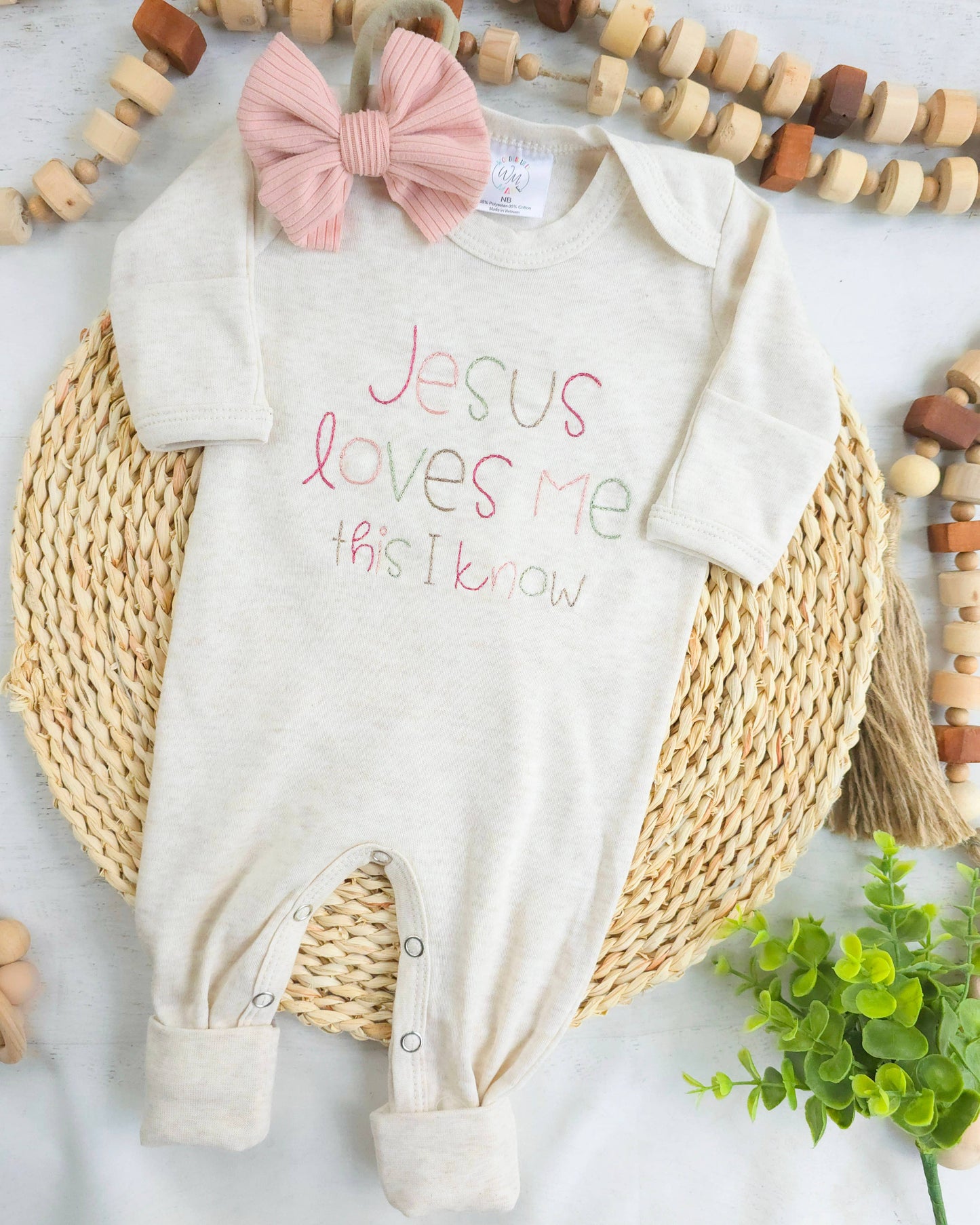 Baby onesie with 'Jesus loves me this I know' text and pink bow on a textured surface.