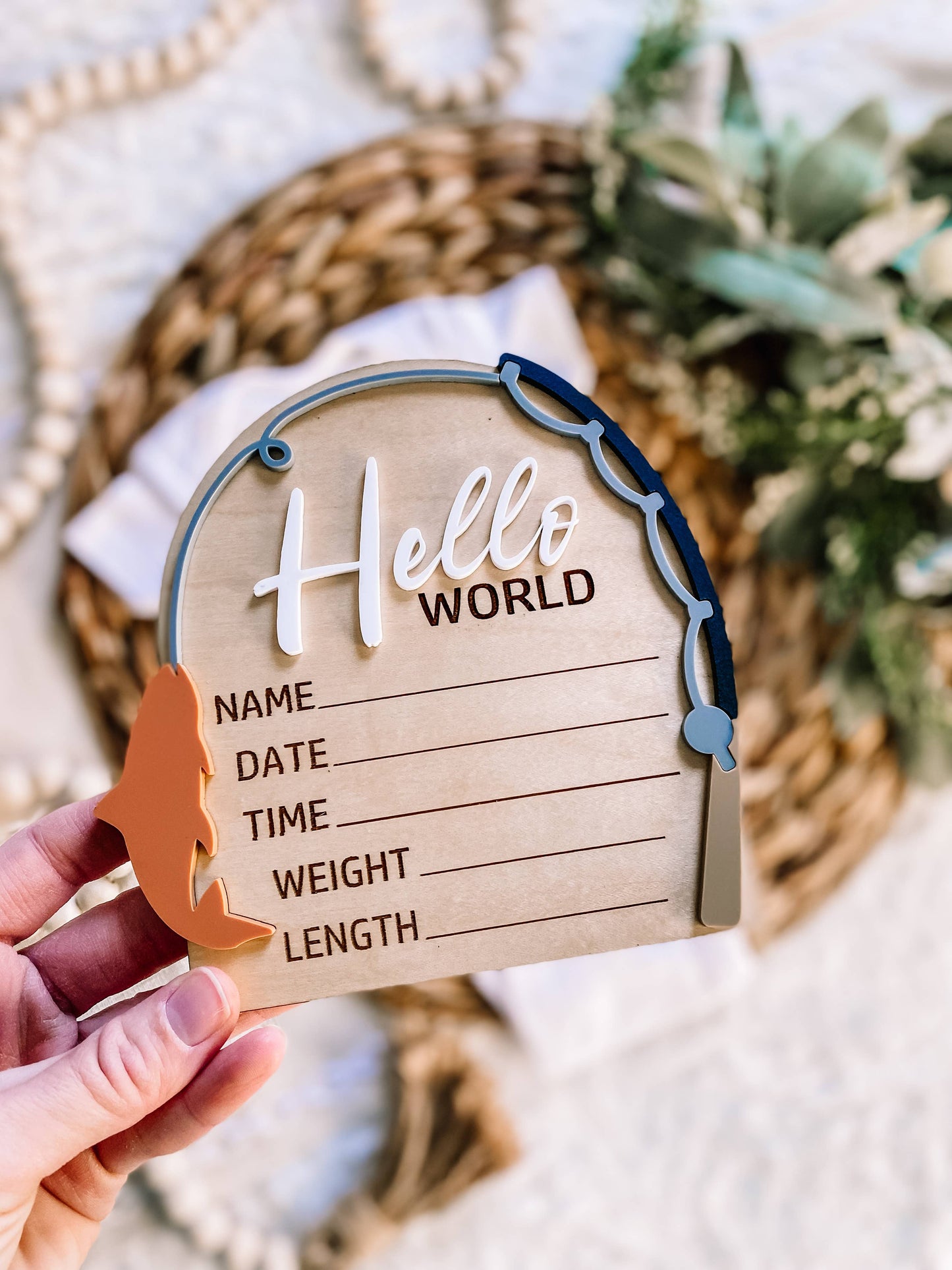 Maple wood “Hello World” birth announcement sign with 3D acrylic lettering and olive fish accent, held in hand with woven mat and greenery background.