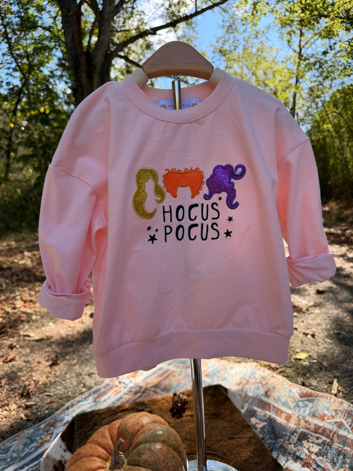 A light pink crewneck sweatshirt with a 'Hocus Pocus' graphic featuring glitter-style orange, purple, and gold script letters, displayed on a wooden hanger on an outdoor stand.