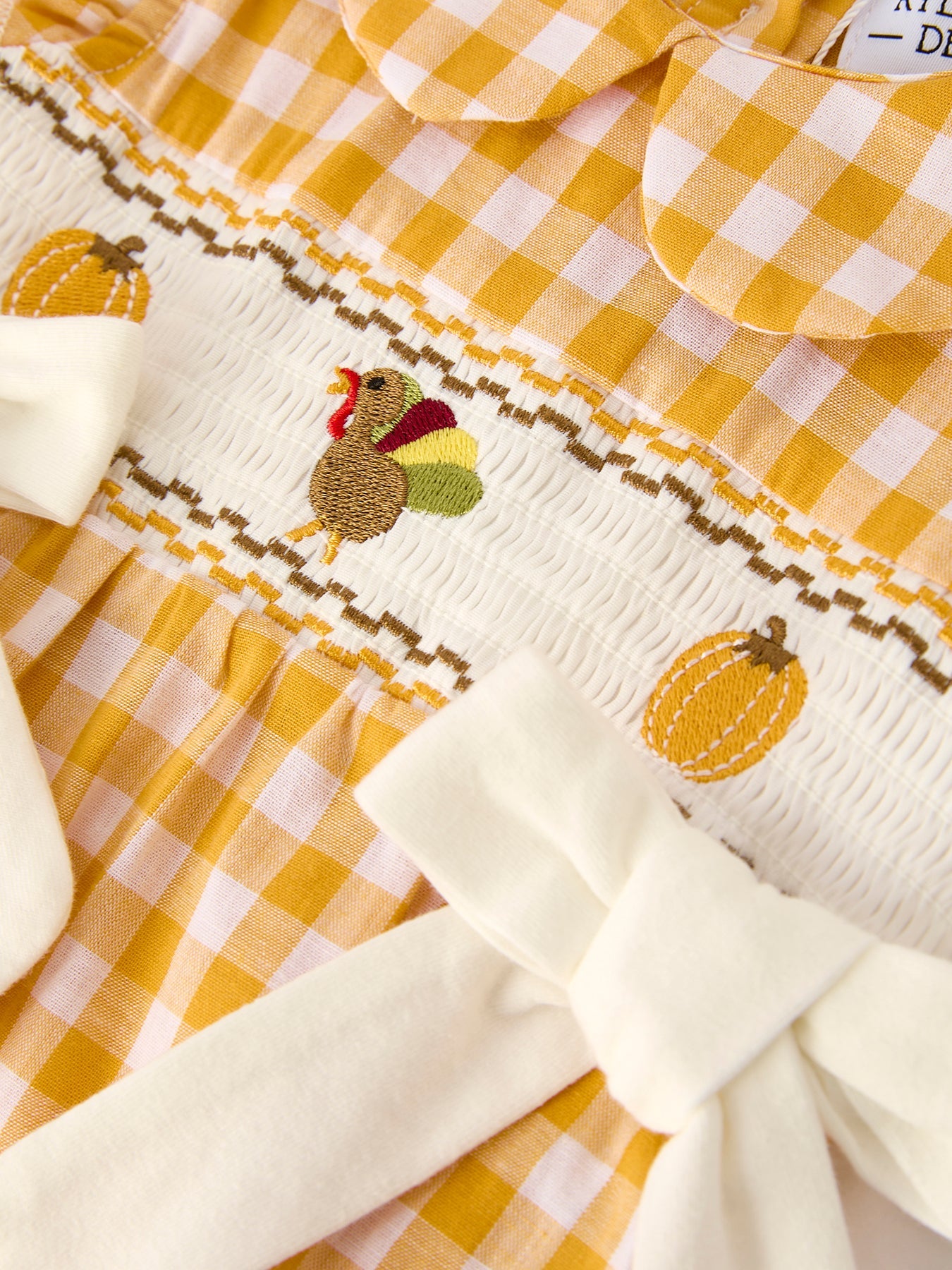 Baby girls yellow gingham bubble romper with pumpkin and turkey smocking, ruffle sleeves, Peter Pan collar, and bow accents.