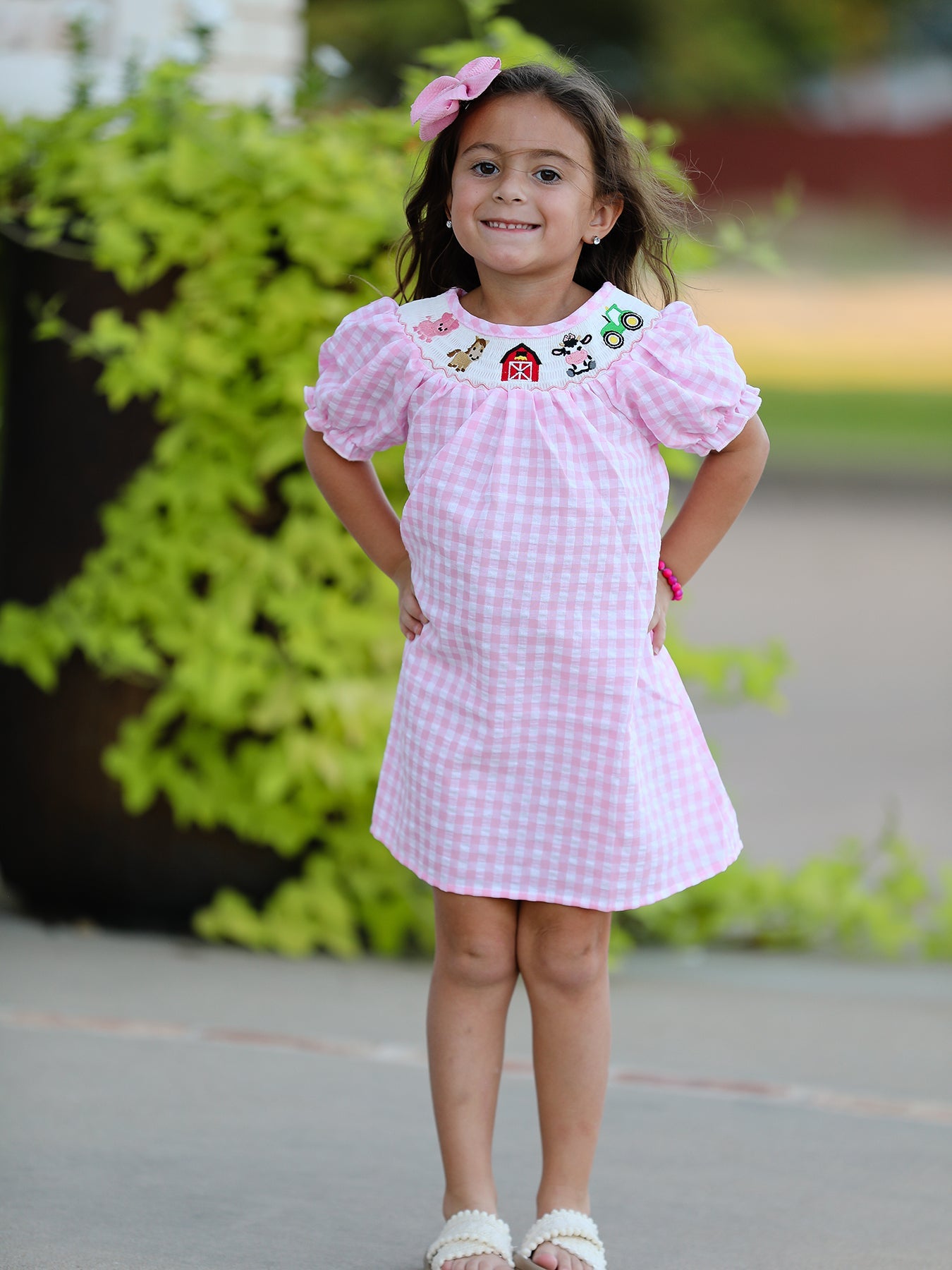 Young girl wearing a pink checkered dress with embroidery, standing outdoors.