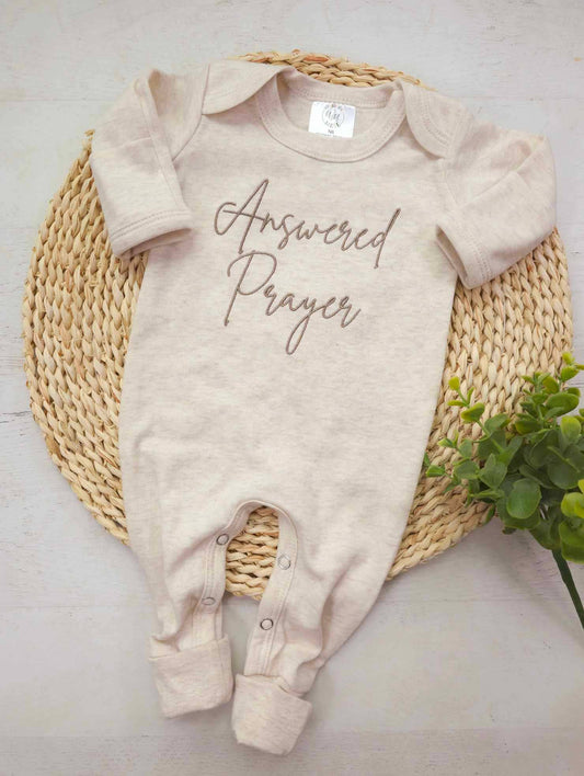 Neutral beige baby romper with brown embroidered “Answered Prayer” text, displayed flat on woven basket mat with greenery accent.