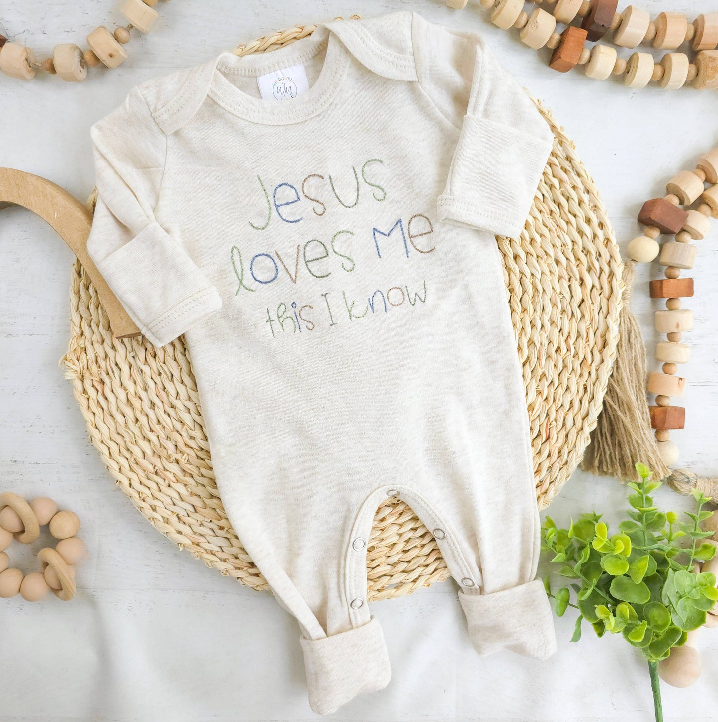 Baby onesie with 'Jesus loves me, this I know' text on a woven mat with wooden toys and greenery.