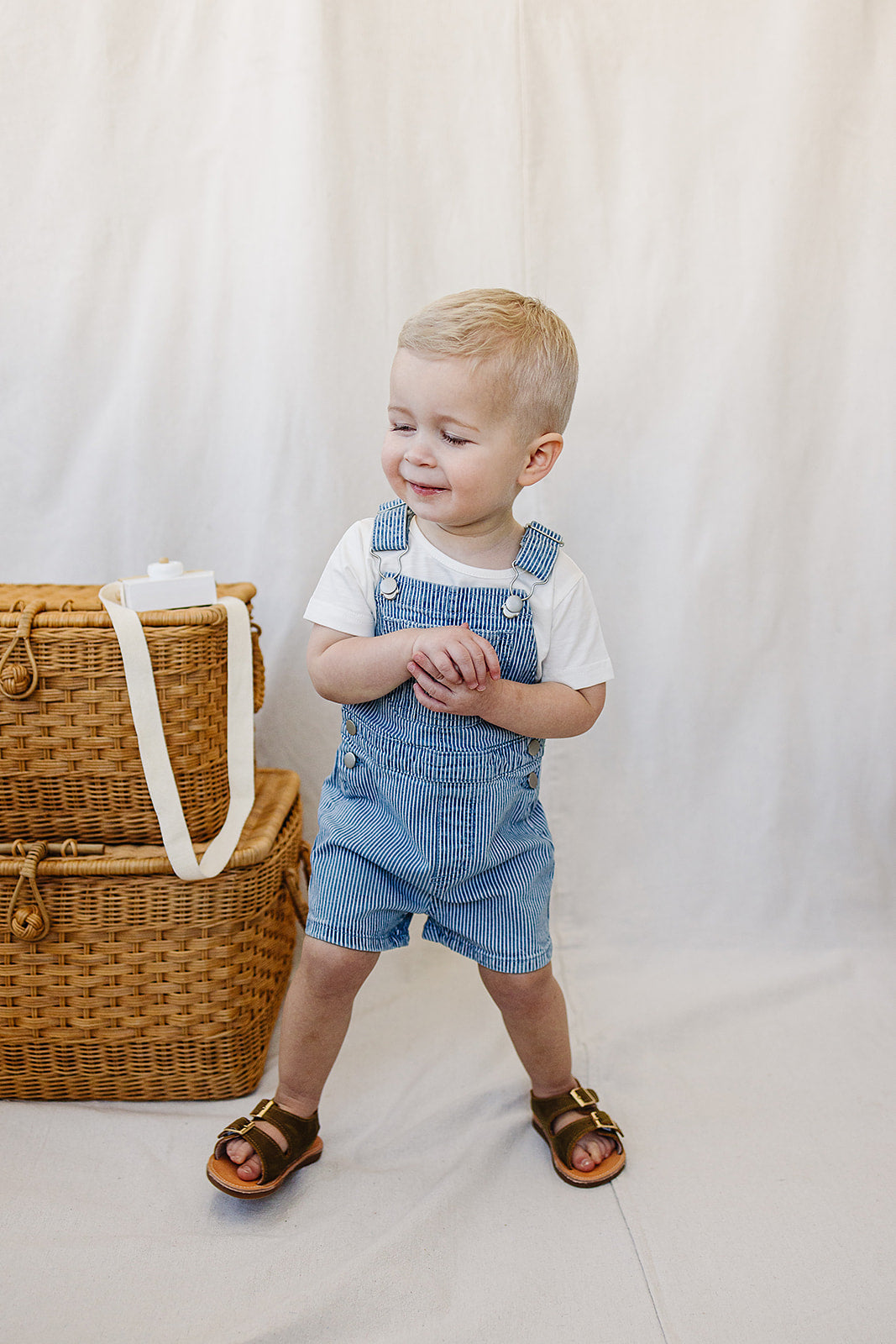 Child wearing a blue denim overalls standing next to a wicker basket on a plain background