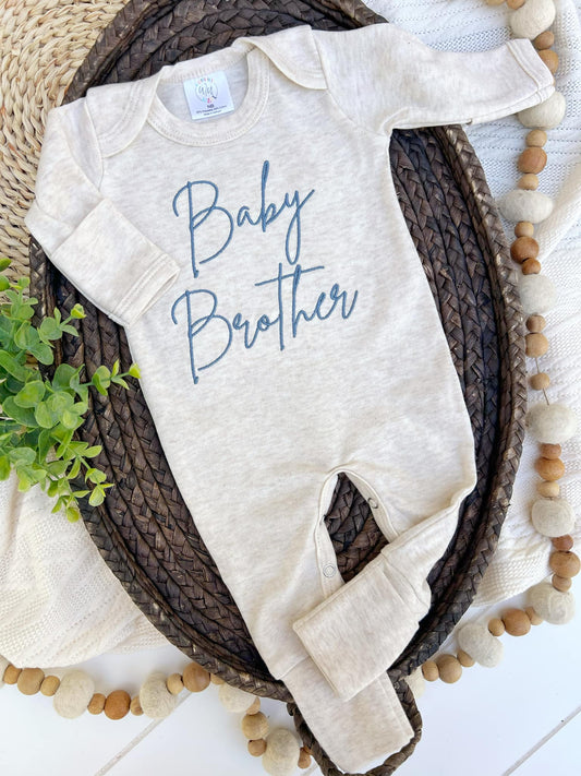 Beige baby onesie with 'Baby Brother' text on a woven tray with decorative elements.