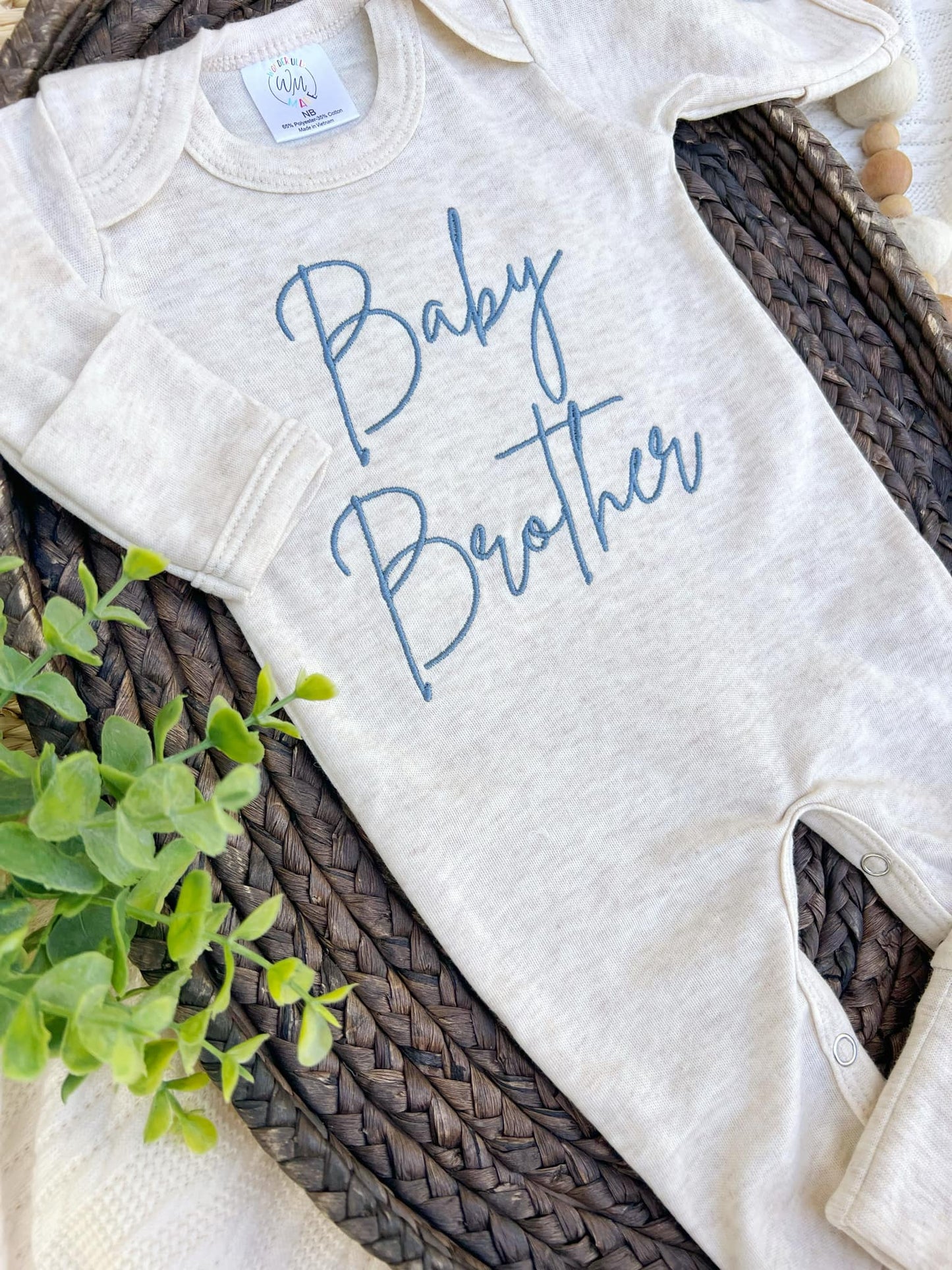 Beige baby onesie with 'Baby Brother' text on a woven surface with greenery.