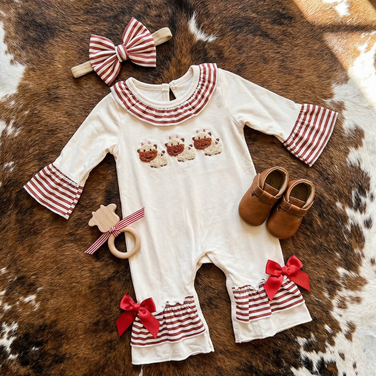 Baby romper with red and white details, smocked highland cows, headband, shoes, and teether on a brown and white cowhide background