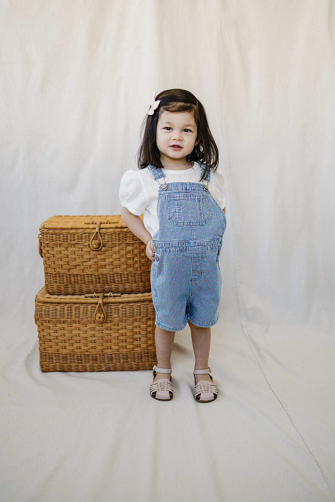 Child wearing denim overalls standing next to wicker baskets on a plain background