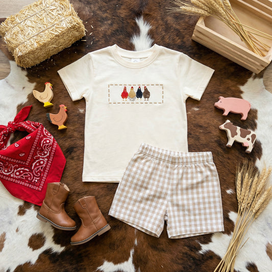 Flat lay toddler boys farm outfit with embroidered chicken shirt and beige gingham shorts styled with boots and farm toys
