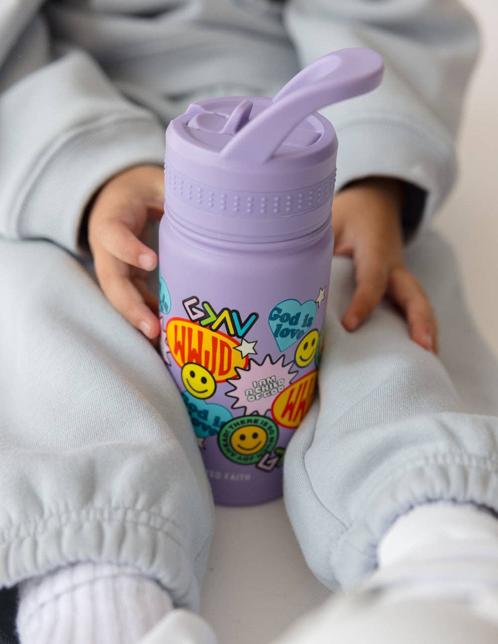 Child holding a purple water bottle with colorful designs and text. BPA Free, faith inspired. 