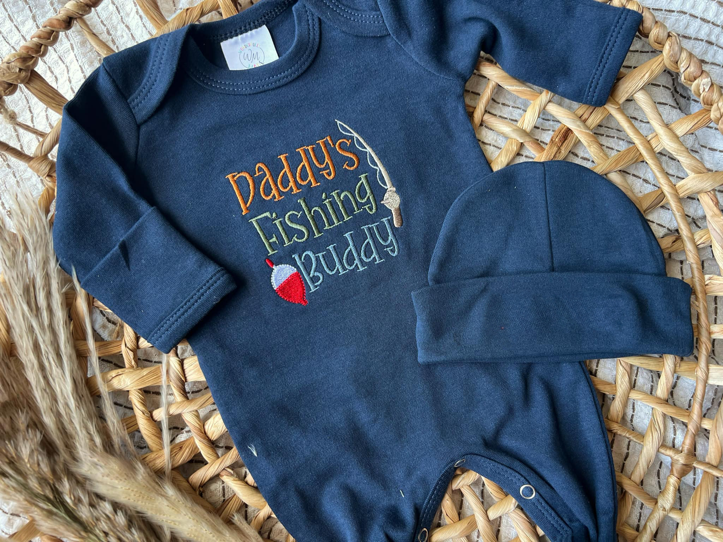 Navy blue baby onesie with 'Daddy's Fishing Buddy' text and matching hat on a woven surface