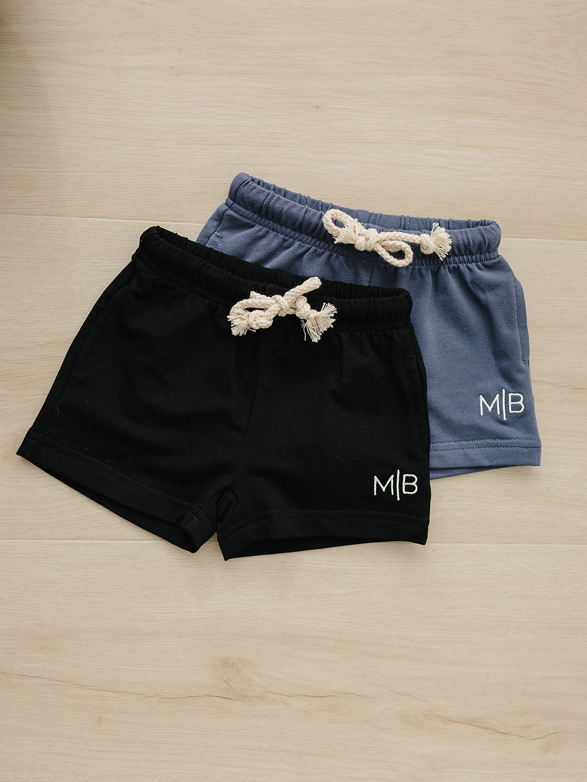 Two pairs of shorts, one black and one blue, with 'MB' logo on a wooden surface.