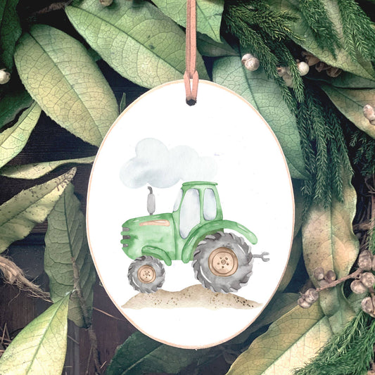 Oval wooden ornament with green tractor illustration and leather hanging cord, displayed on greenery background.