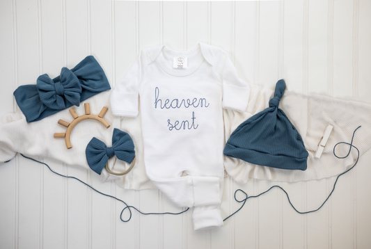 White baby romper with light blue “heaven sent” embroidery, displayed flat with neutral baby accessories on a white background.