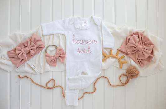White baby romper with pink “heaven sent” embroidery, styled with blush bow and neutral baby accessories on a white background.