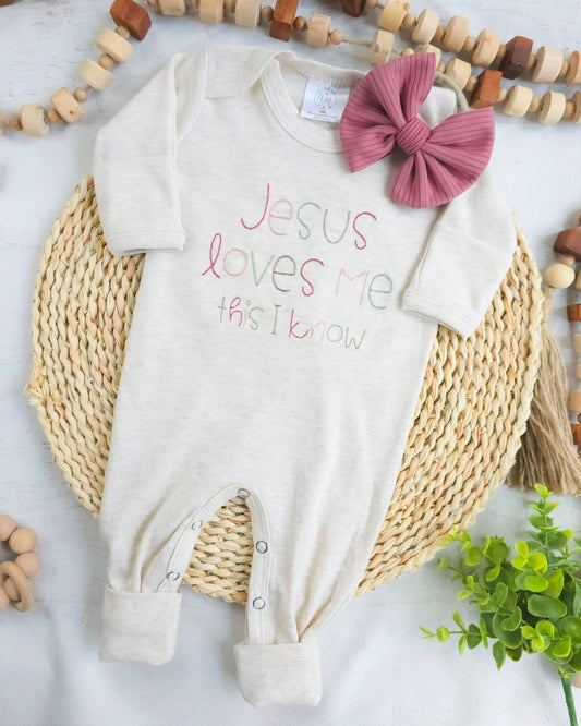 Baby onesie with 'Jesus loves me this I know' text and a matching bow on a woven mat.