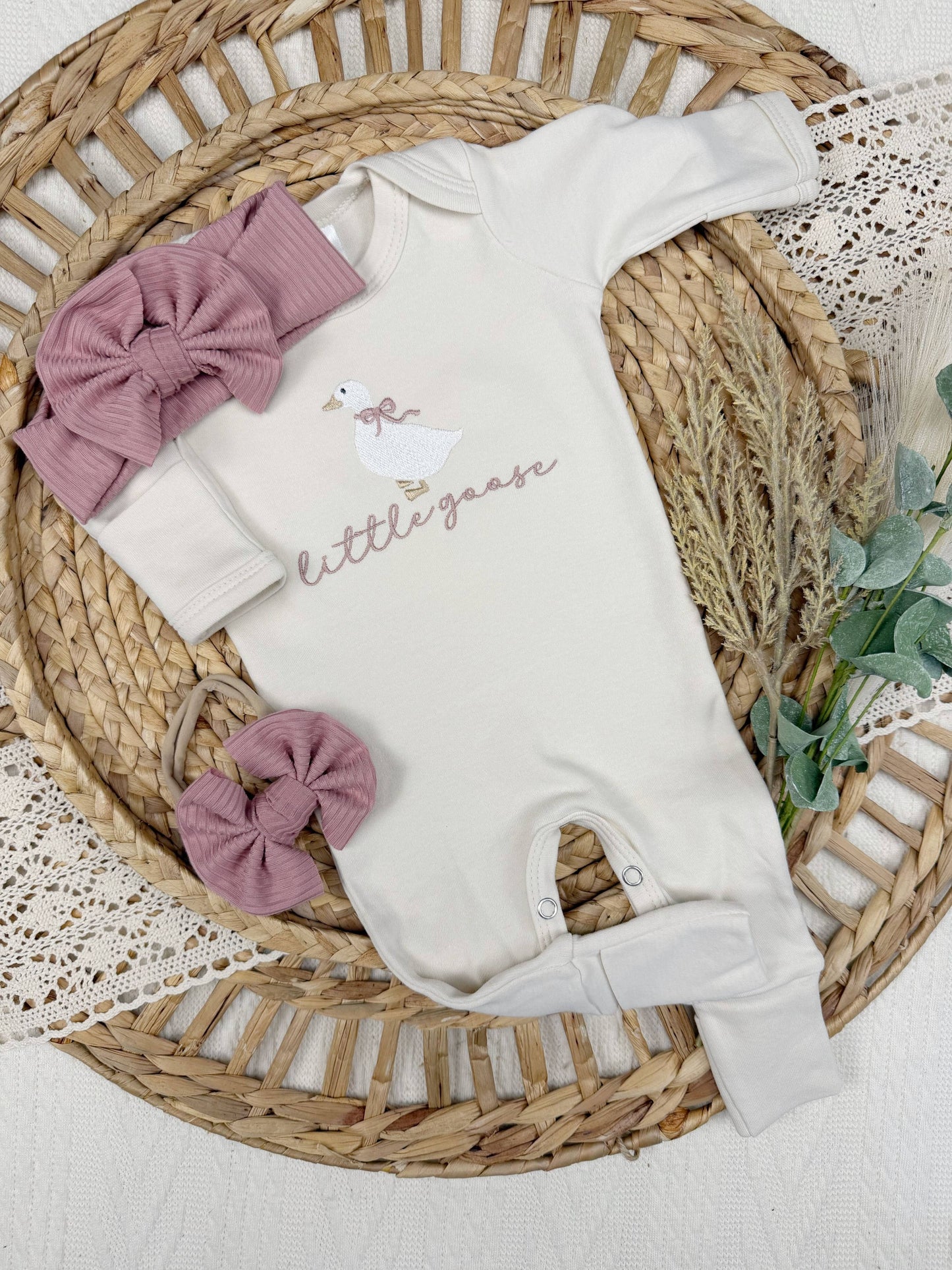 cream baby onesie with 'little goose' text, pink headbands, and a ring on a woven basket.