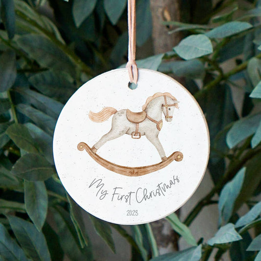 Round wooden ornament with rocking horse design and text “My First Christmas 2025,” hanging on leather cord in front of green leaves.