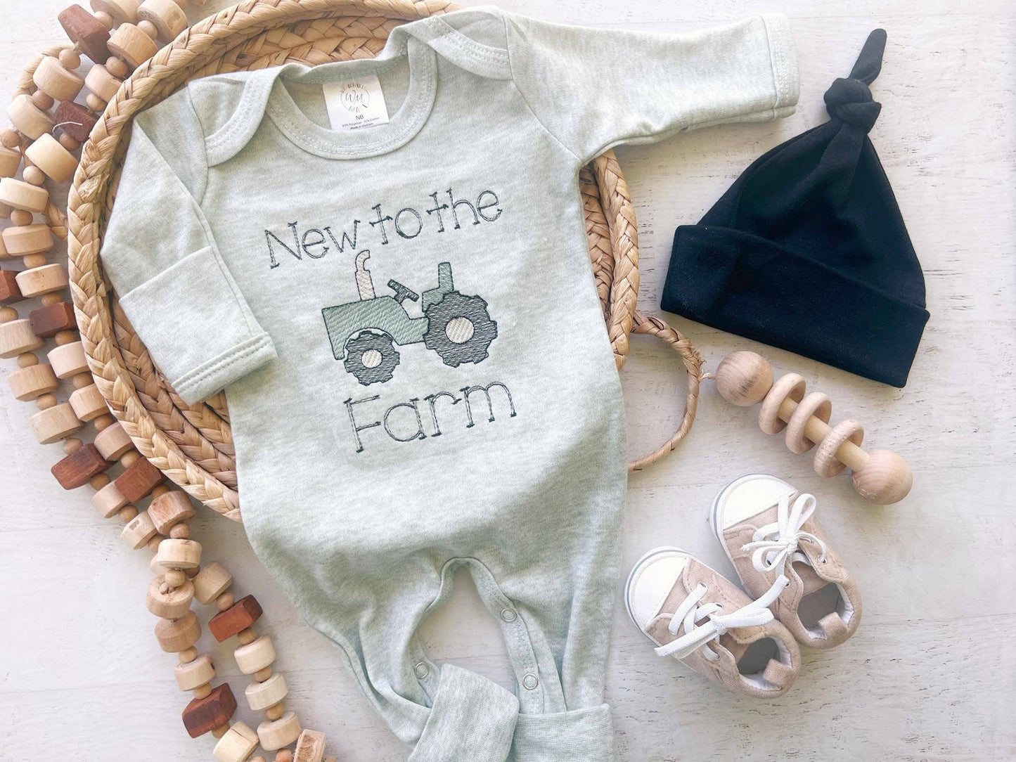 Baby onesie with 'New to the Farm' text and tractor graphic, surrounded by baby items on a light surface.