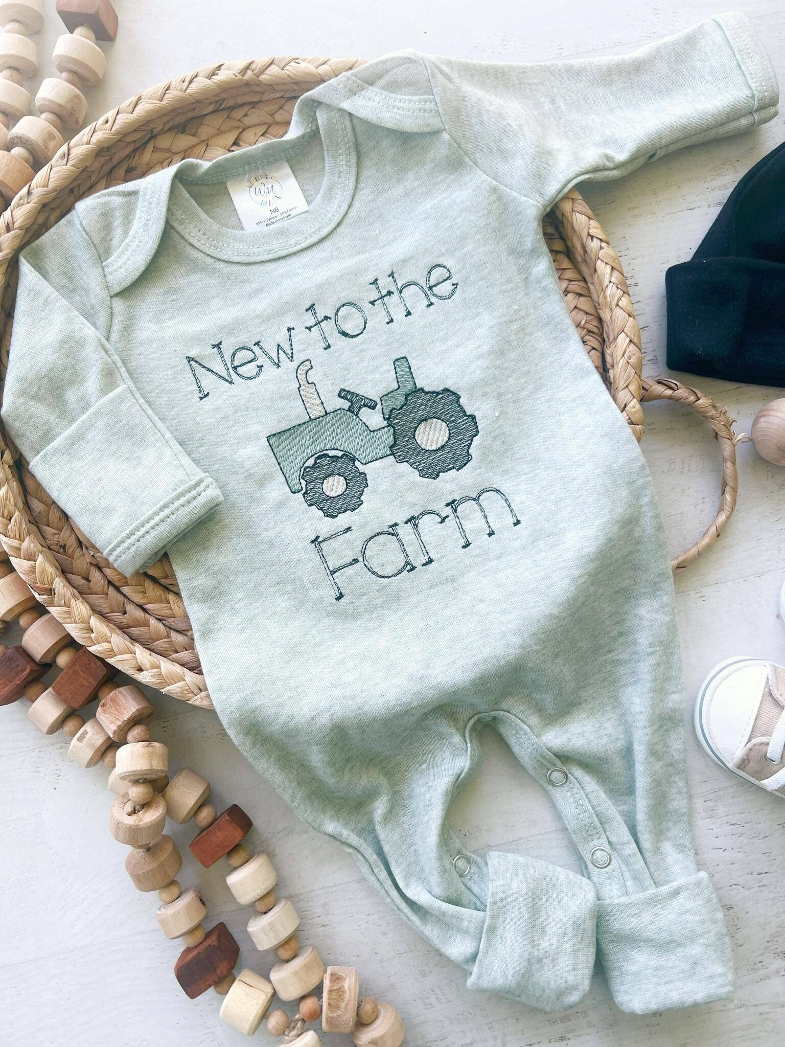 Sage green baby romper with black embroidered “New to the Farm” text and tractor design, displayed flat with black knotted hat beside it.
