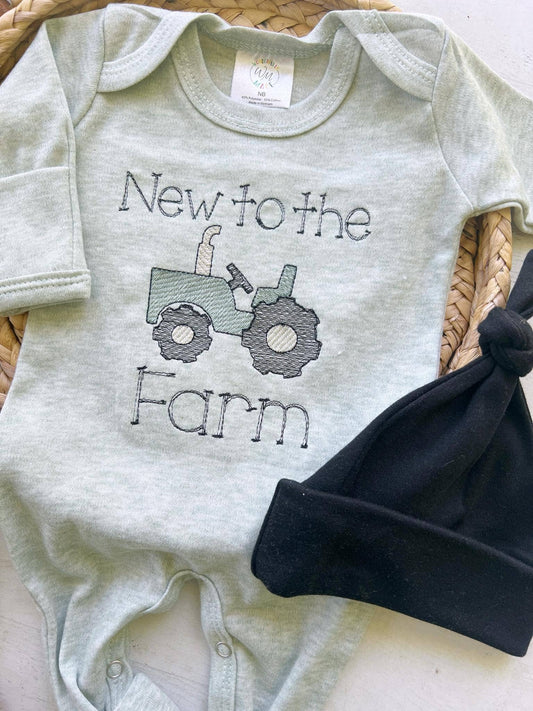 Sage green baby romper with black embroidered “New to the Farm” text and tractor design, displayed flat with black knotted hat beside it.