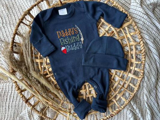 Navy blue baby onesie with 'Daddy's Fishing Buddy' text and matching hat on a woven mat.