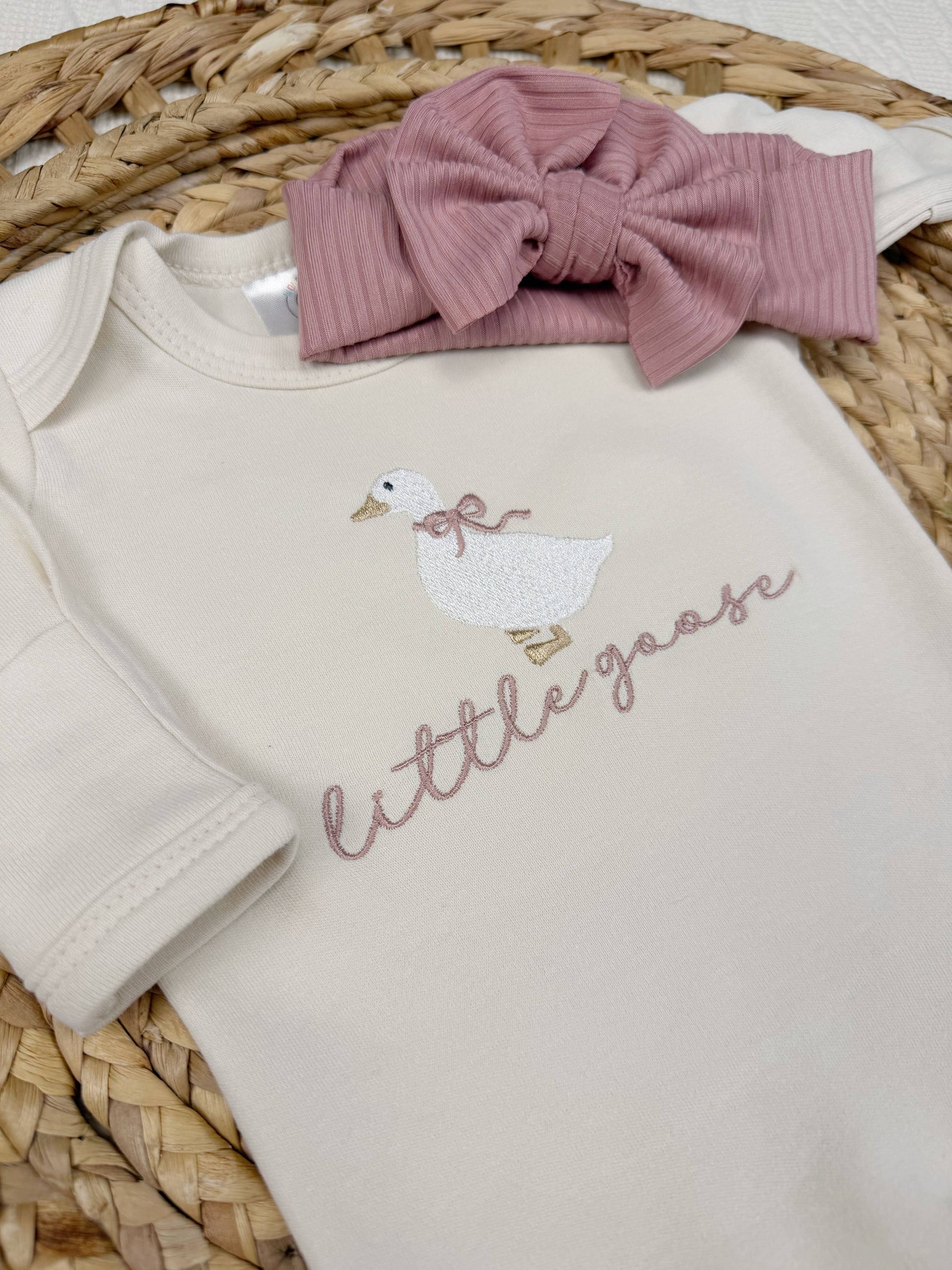 Beige baby onesie with duck design and 'little goose' text, paired with a pink bow headband on a woven surface.