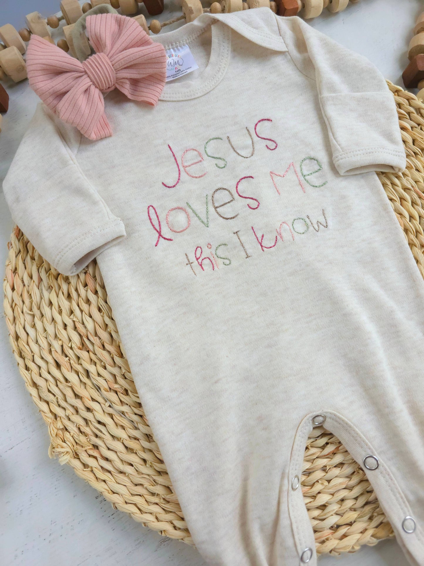 Beige baby romper with 'Jesus loves me this I know' text and pink bow on a woven mat.
