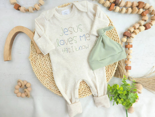 Baby onesie with 'Jesus loves me, has I know' text, green hat, and wooden toys on a light background