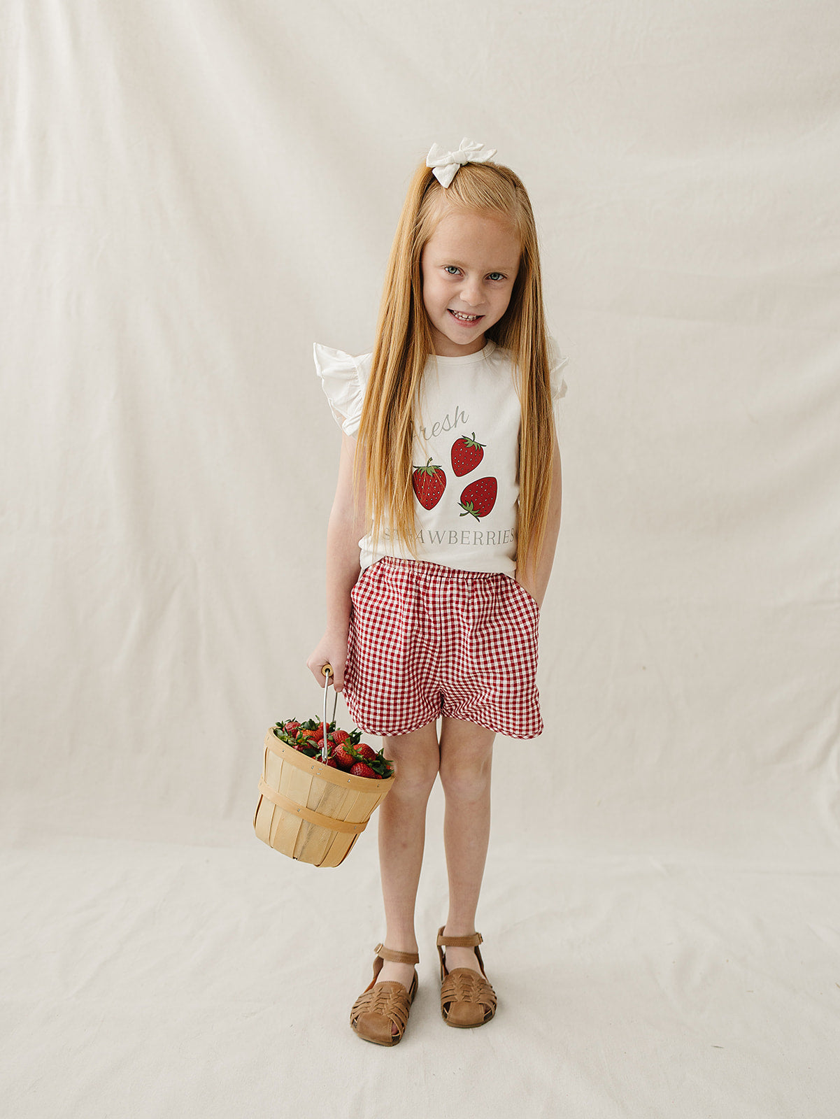 Young girl in a strawberry-themed outfit holding a basket of strawberries on a plain background