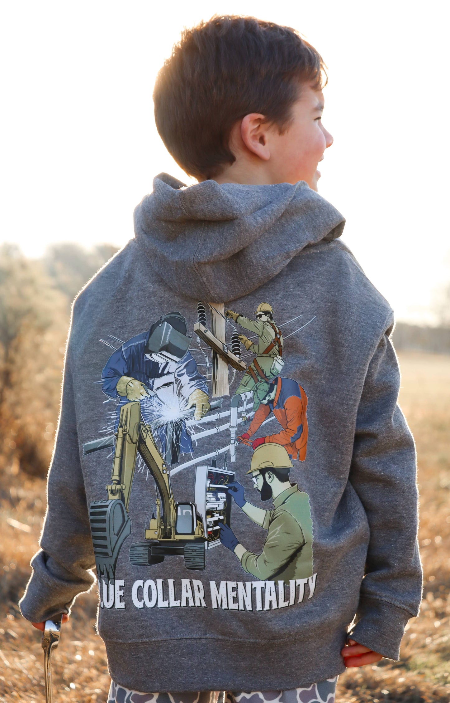 Child wearing Southern Hyde Blue Collar Mentality gray kids hoodie with trades graphic on the back outdoors