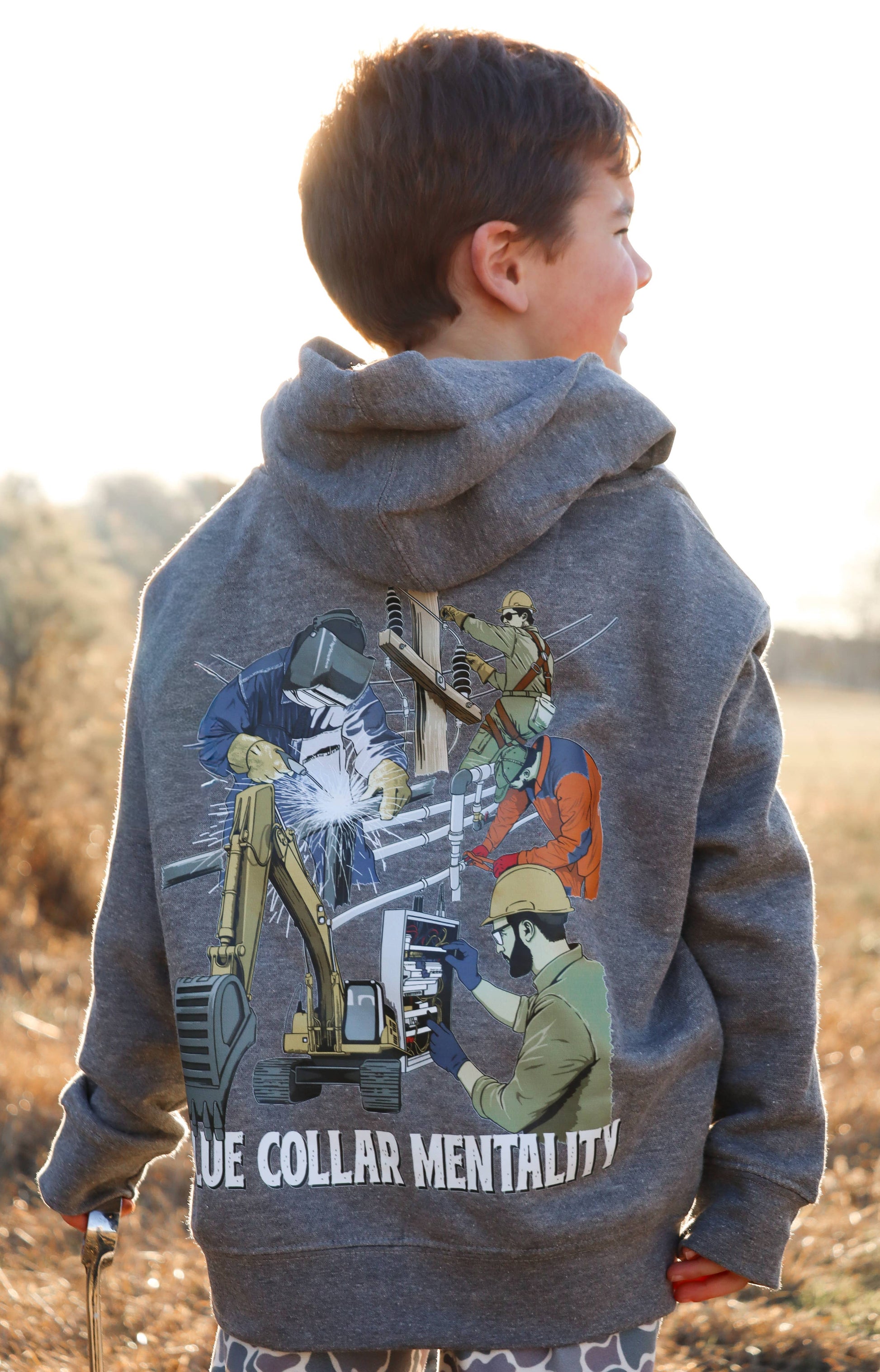 Child wearing Southern Hyde Blue Collar Mentality gray kids hoodie with trades graphic on the back outdoors
