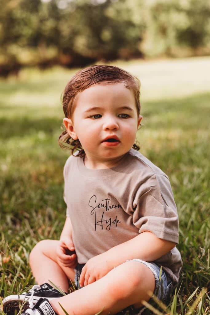 Toddler wearing Southern Hyde Co neutral tan graphic tee sitting in grass outdoor lifestyle photo