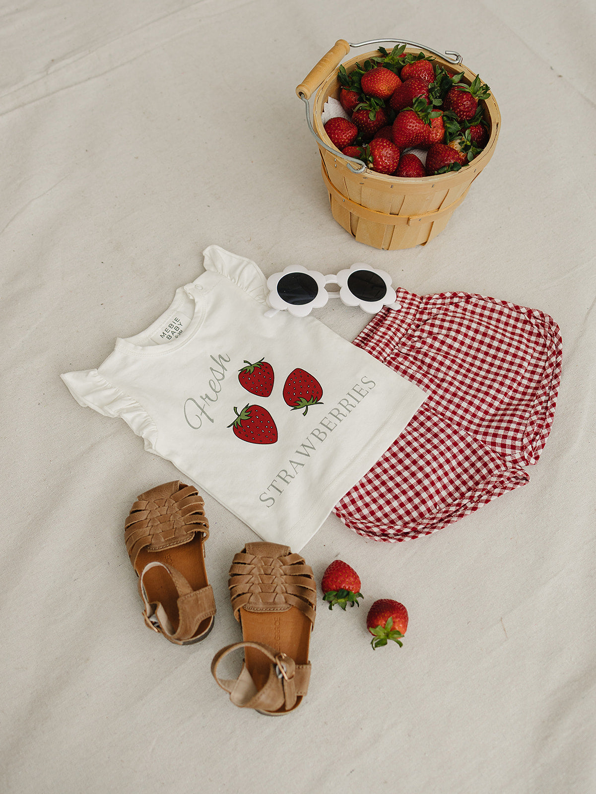 Baby outfit with 'Good Vibes Strawberry' shirt, sandals, and a basket of strawberries on a neutral background