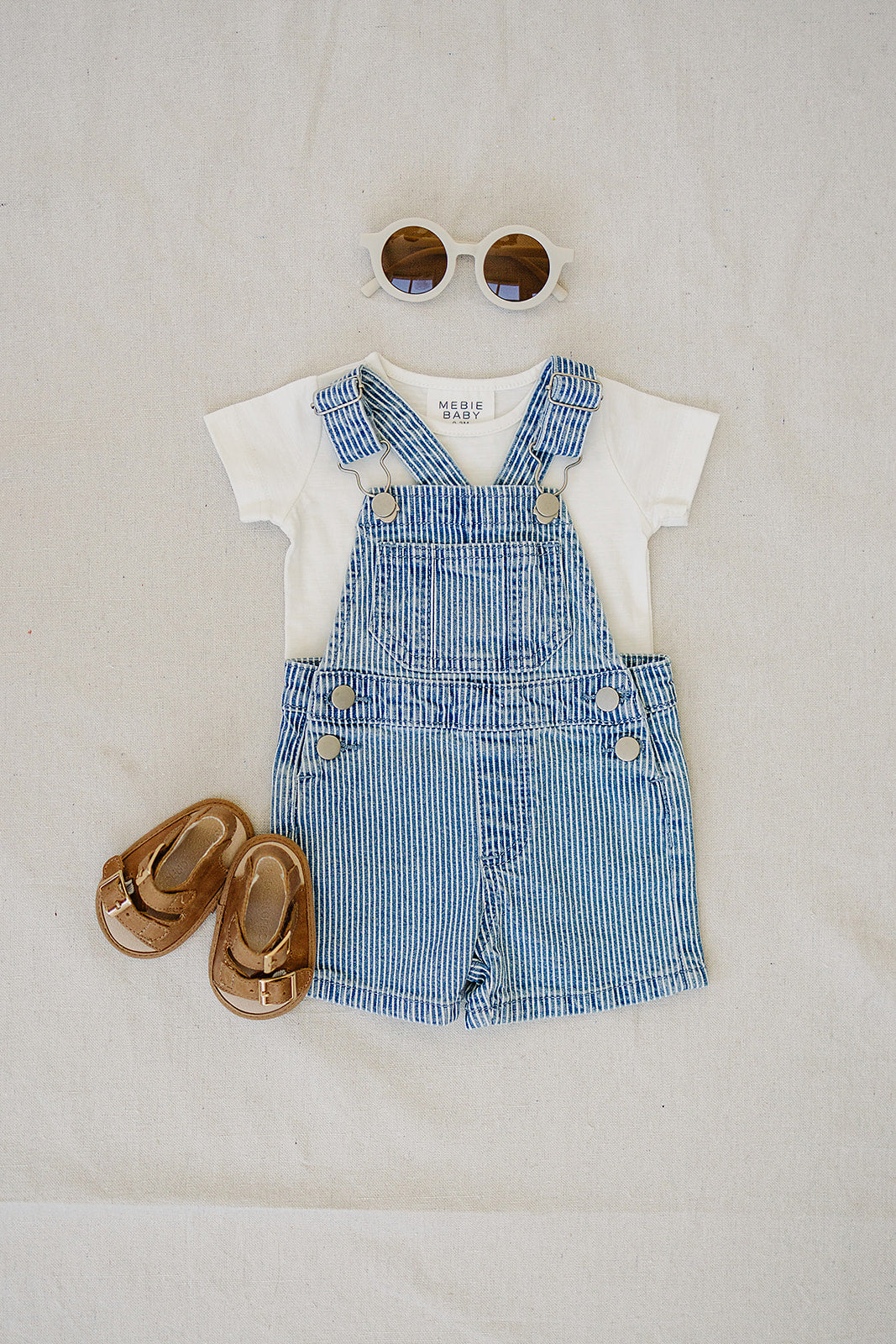 Children's outfit with denim overalls, white shirt, sandals, and sunglasses on a light background