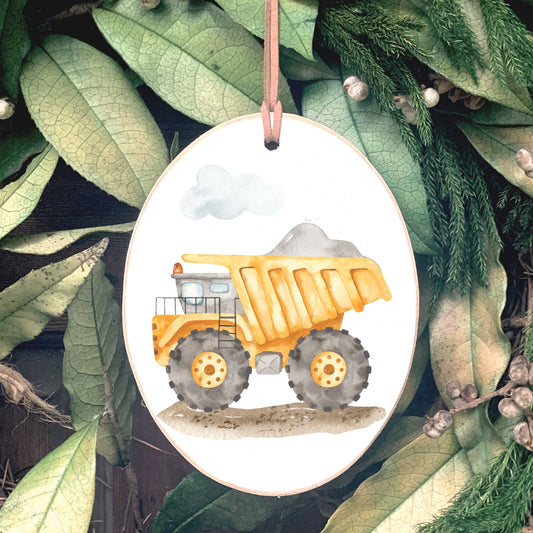 Oval wooden ornament with yellow dump truck illustration and leather hanging cord, displayed on greenery background.